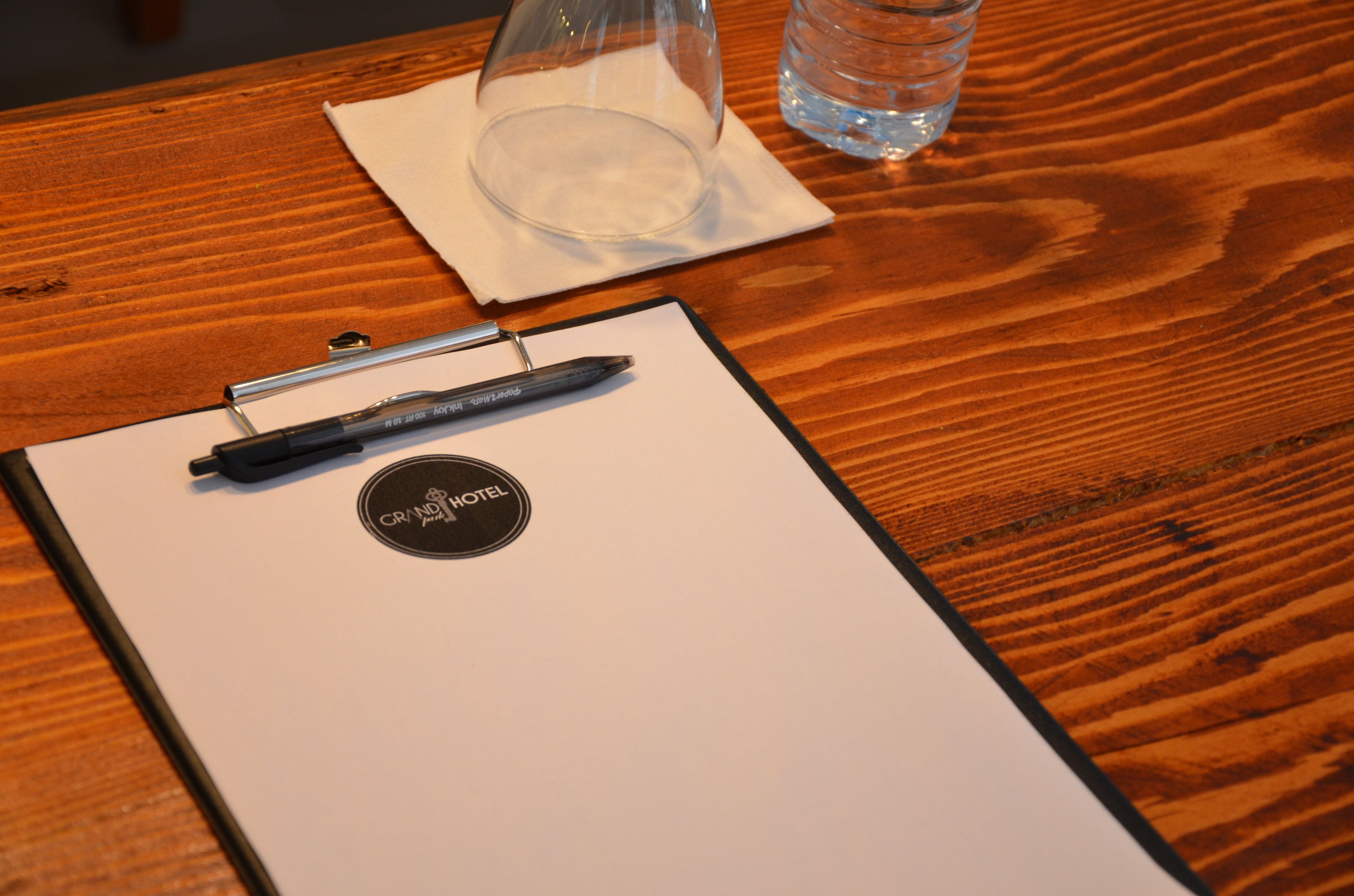 A wooden table with a clipboard, a pen, a glass, and a water bottle.