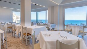 A restaurant with white tables and chairs, large windows offering a view of the sea, and a modern interior design. - Hotel Peschici (Peschici)