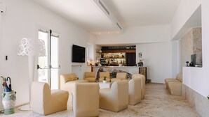 A modern living room with beige sofas arranged around a central coffee table, a kitchen area with a bar visible in the background, and a large window with decorative curtains. - Hotel Peschici (Peschici)