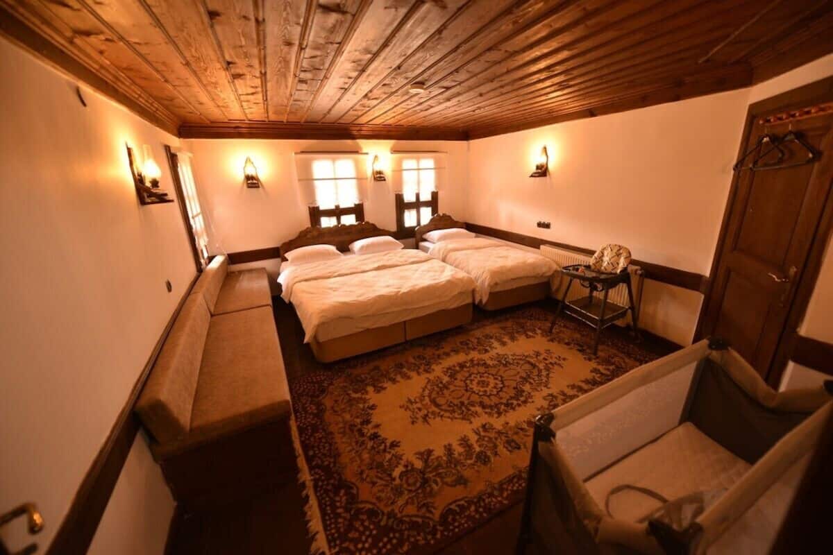 A room with a wooden ceiling, two beds with white linens, a patterned rug, and wall-mounted lights.