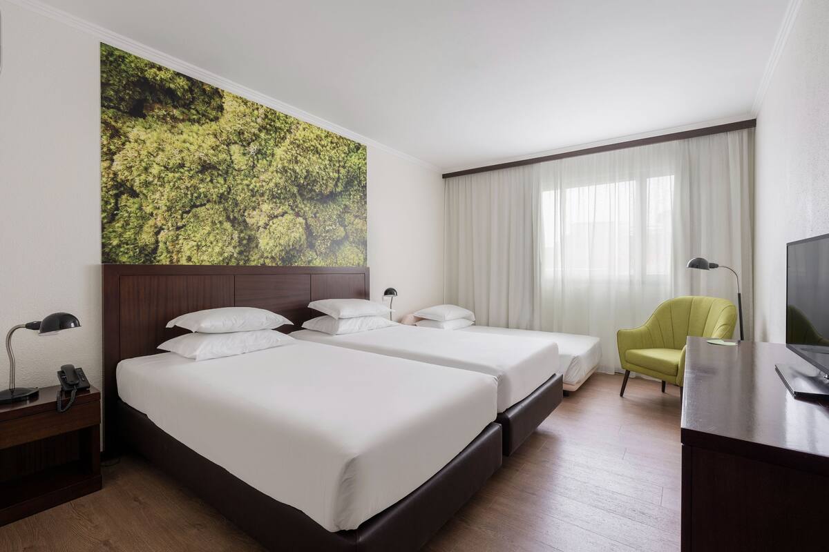 A hotel room with two beds, a desk, a chair, a TV, and a large window with curtains.