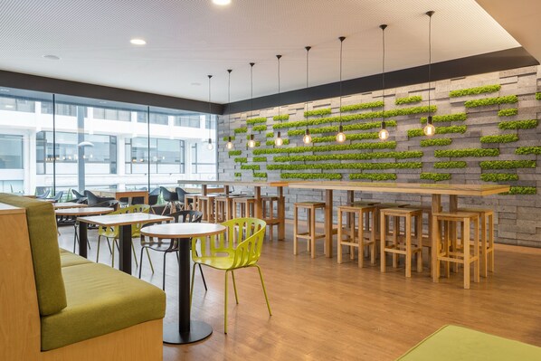 A modern cafe with wooden tables and chairs, a green cushioned bench, and a wall with vertical gardens. - Neat Hotel Avenida (Ponta Delgada)