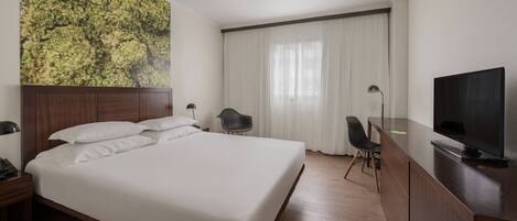 Quarto de hotel com uma cama grande, uma escrivaninha com televisão, uma cadeira e uma luminária de mesa.