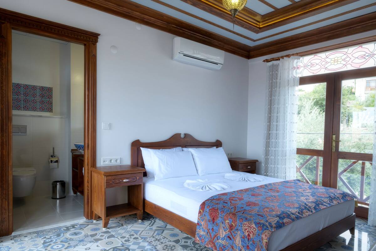 A bedroom with a wooden bed, white bedding, a patterned throw, a nightstand, an air conditioning unit, and a view of greenery through a window.