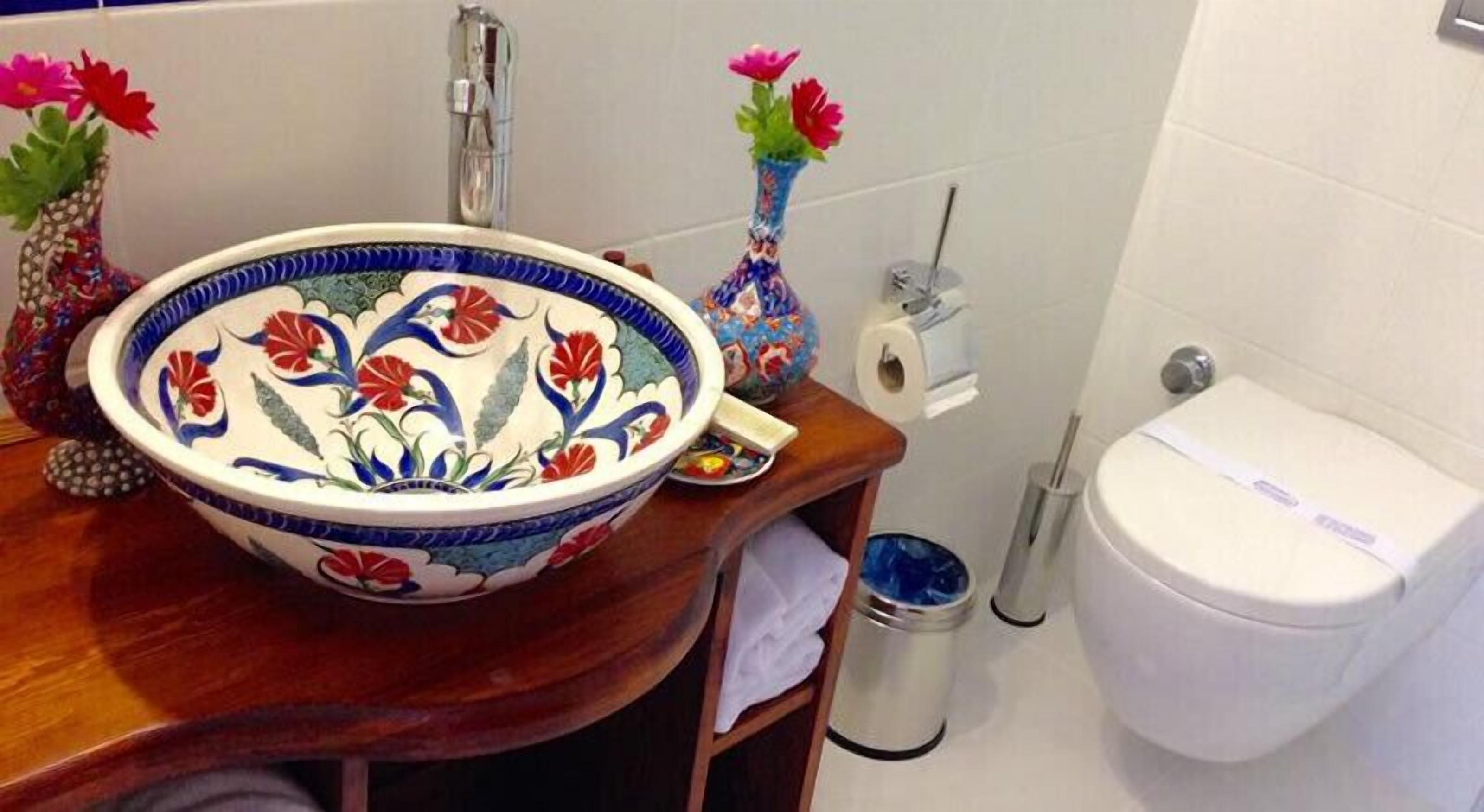 Ein Badezimmer mit einem großen, dekorativen Waschbecken, zwei Vasen mit Blumen, einer Toilette und einem Mülleimer.