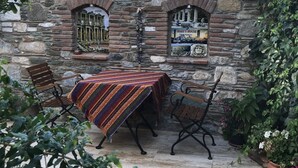 Una pared de piedra con dos ventanas arqueadas, una mesa con mantel a rayas y varias sillas.