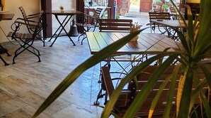 Un espacio interior amplio con suelo de madera, sillas de estructura metálica y mesas. Se aprecia una ventana grande con vistas al exterior, y un ventilador a la izquierda.