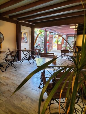 Un espacio interior amplio con suelo de madera, sillas de estructura metálica y mesas. Se aprecia una ventana grande con vistas al exterior, y un ventilador a la izquierda.