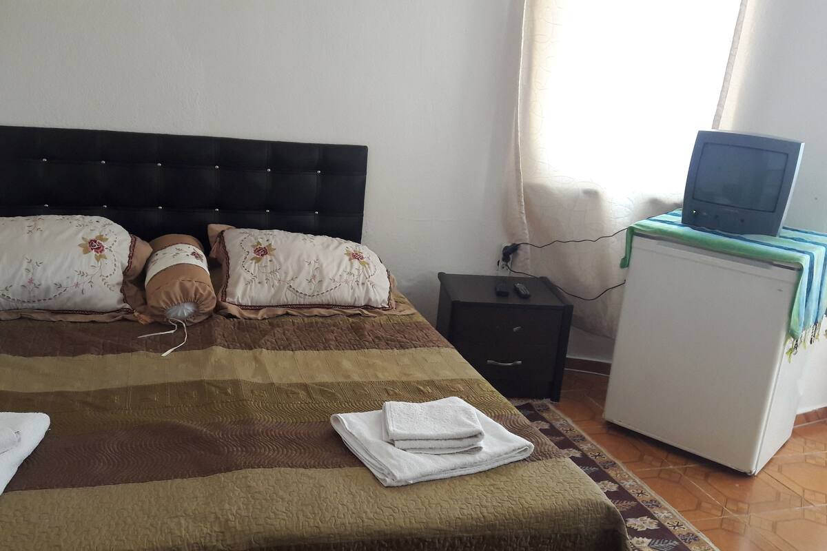Uma cama bem arrumada com almofadas florais, uma pequena mesa de cabeceira com um controle remoto, uma televisão em um suporte e um chão de azulejos.