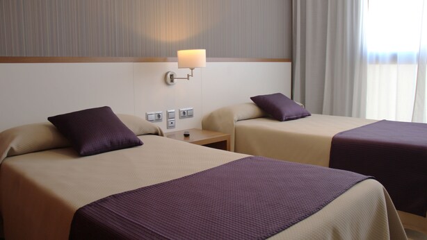 A hotel room with two beds, each with a purple pillow and a beige bedspread. There is a wall-mounted lamp on the left side of the image, and a window with curtains on the right. - Hotel Teruel (Vinaros)