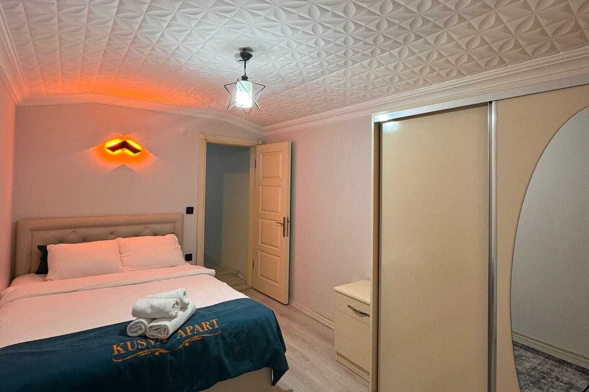 A hotel room with a geometric patterned ceiling, a bed with a blue bedspread, a wardrobe, and a small bedside table.