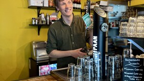 A barista at a coffee shop counter with a menu board displaying various drinks and prices. - The Golden Stork - Hostel (The Hague)