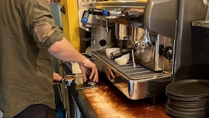 A person operating a coffee machine in a kitchen area with a yellow door and stainless steel appliances. - The Golden Stork - Hostel (The Hague)