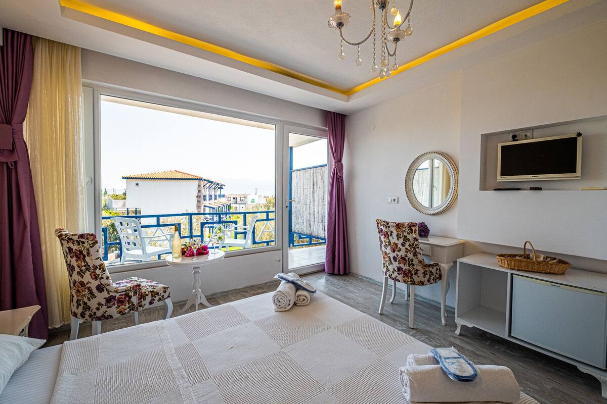Een hotelkamer met een bed, twee stoelen, een kleine tafel met een fles en bloemen, een televisie en uitzicht op zee en gebouwen.
