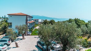 Un hôtel à plusieurs étages, doté d’un toit recouvert de tuiles rouges, d’un parking où sont garées des voitures, et offrant une vue sur la mer et les montagnes en arrière-plan.