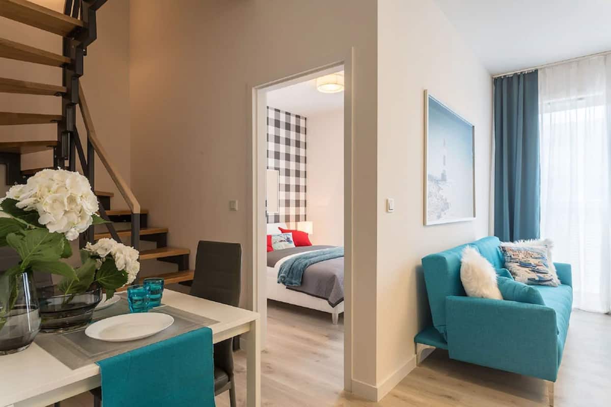 A modern apartment with a staircase, a dining area with a table and chairs, a teal sofa, and a bed with checkered bedding.