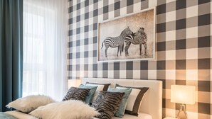 Un dormitorio ordenado con pared a cuadros, una obra de arte con cebra enmarcada, una cama blanca con almohadas mullidas y una lámpara de noche.