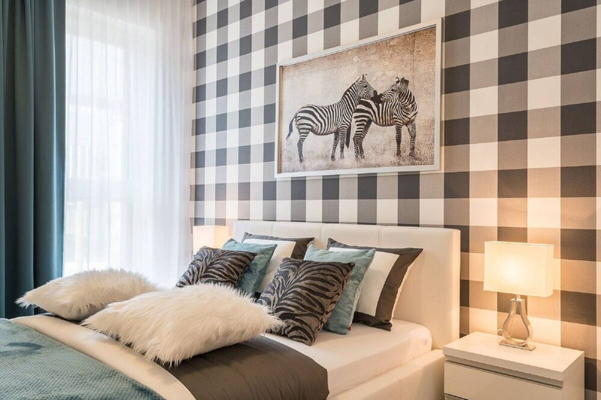 A neatly arranged bedroom with a checkered wall, a framed zebra artwork, a white bed with fluffy pillows, and a bedside lamp.
