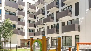 Un parque infantil con una estructura para escalar y un tobogán, junto a un edificio de apartamentos moderno con balcones.