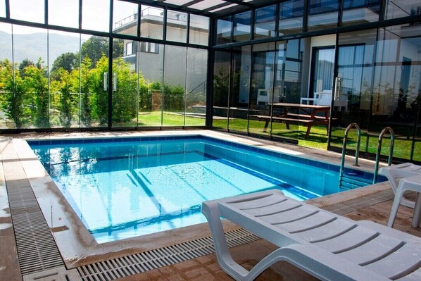A swimming pool with clear blue water, surrounded by a glass enclosure with a view of greenery and buildings.