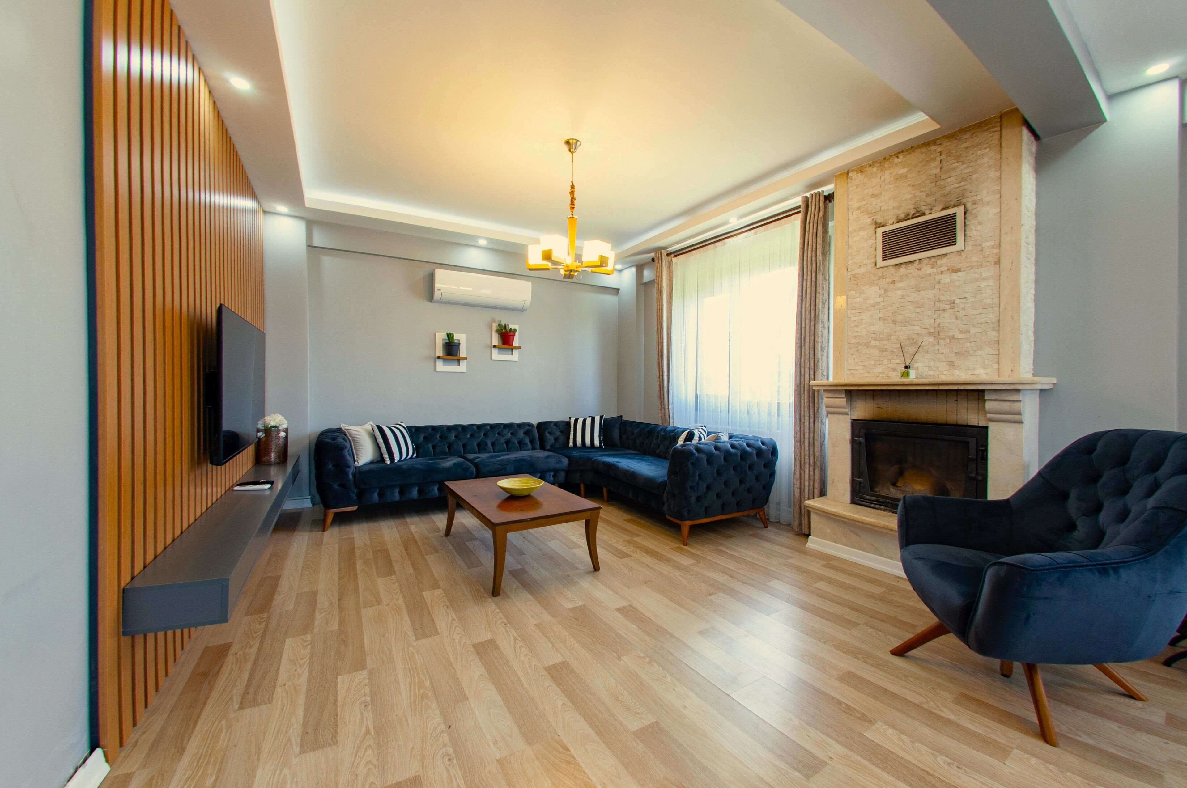 Un salon moderne avec un canapé d’angle, une table basse, un téléviseur fixé au mur, une cheminée et une grande fenêtre avec des rideaux.