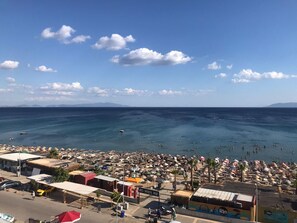 Ein Strand mit Sonnenschirmen und badenden Menschen, eine Bar namens Rio Beach Bar, und ein Blick auf das Meer und ferne Berge.