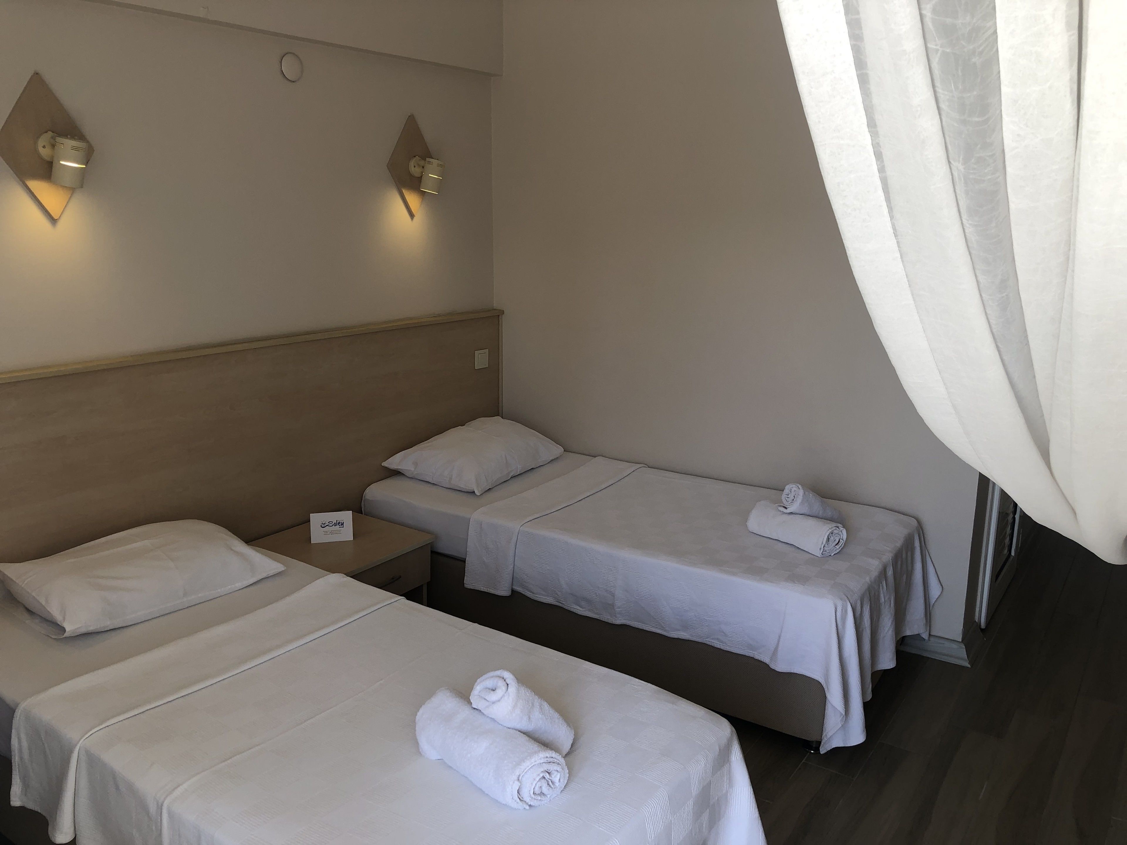 A hotel room with two single beds, a wooden headboard, a nightstand with a sign, and wall-mounted lighting.