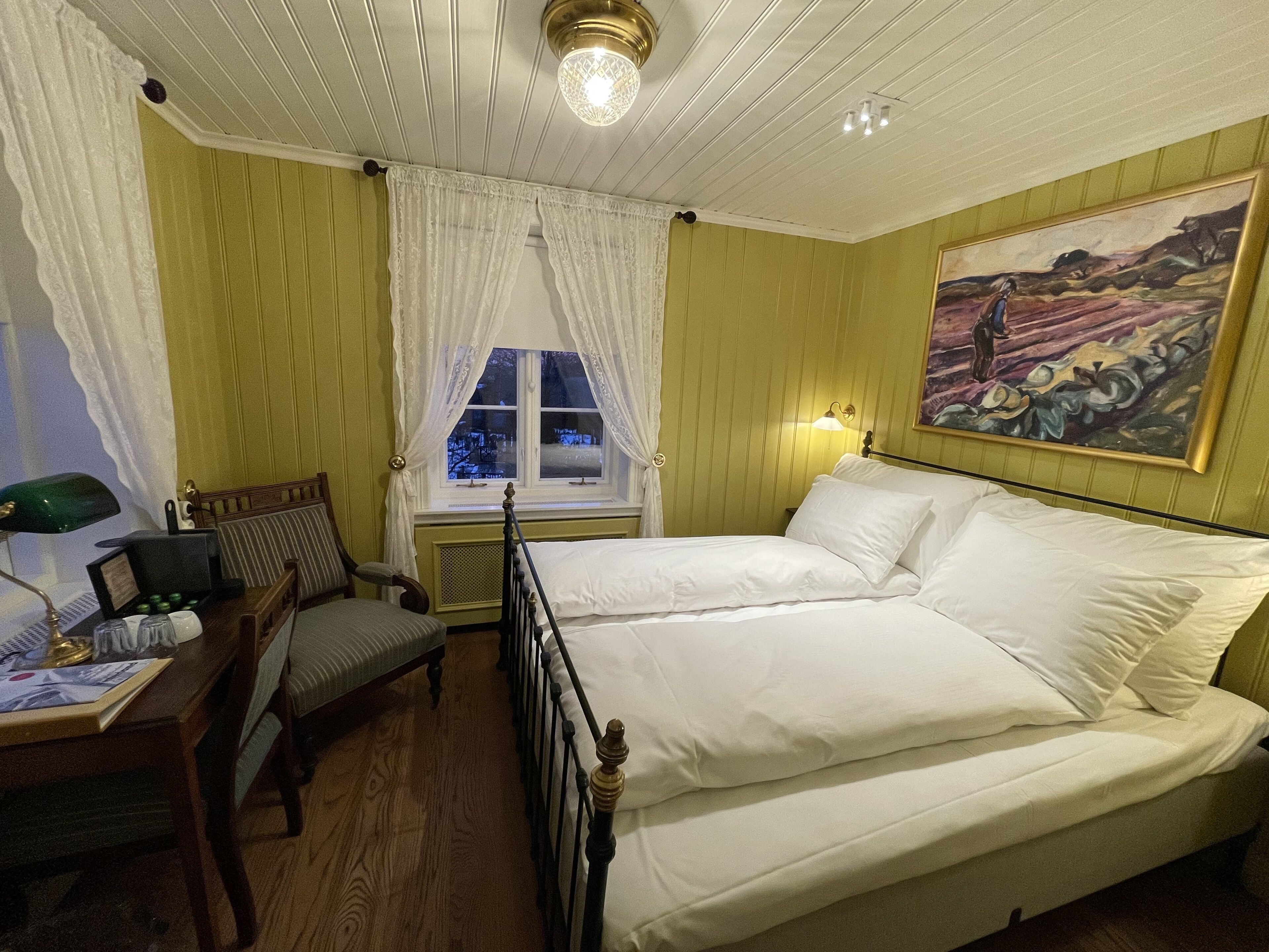 Une chambre d’hôtel avec un lit, un bureau avec une lampe, une chaise, une fenêtre avec des rideaux et un tableau au mur.
