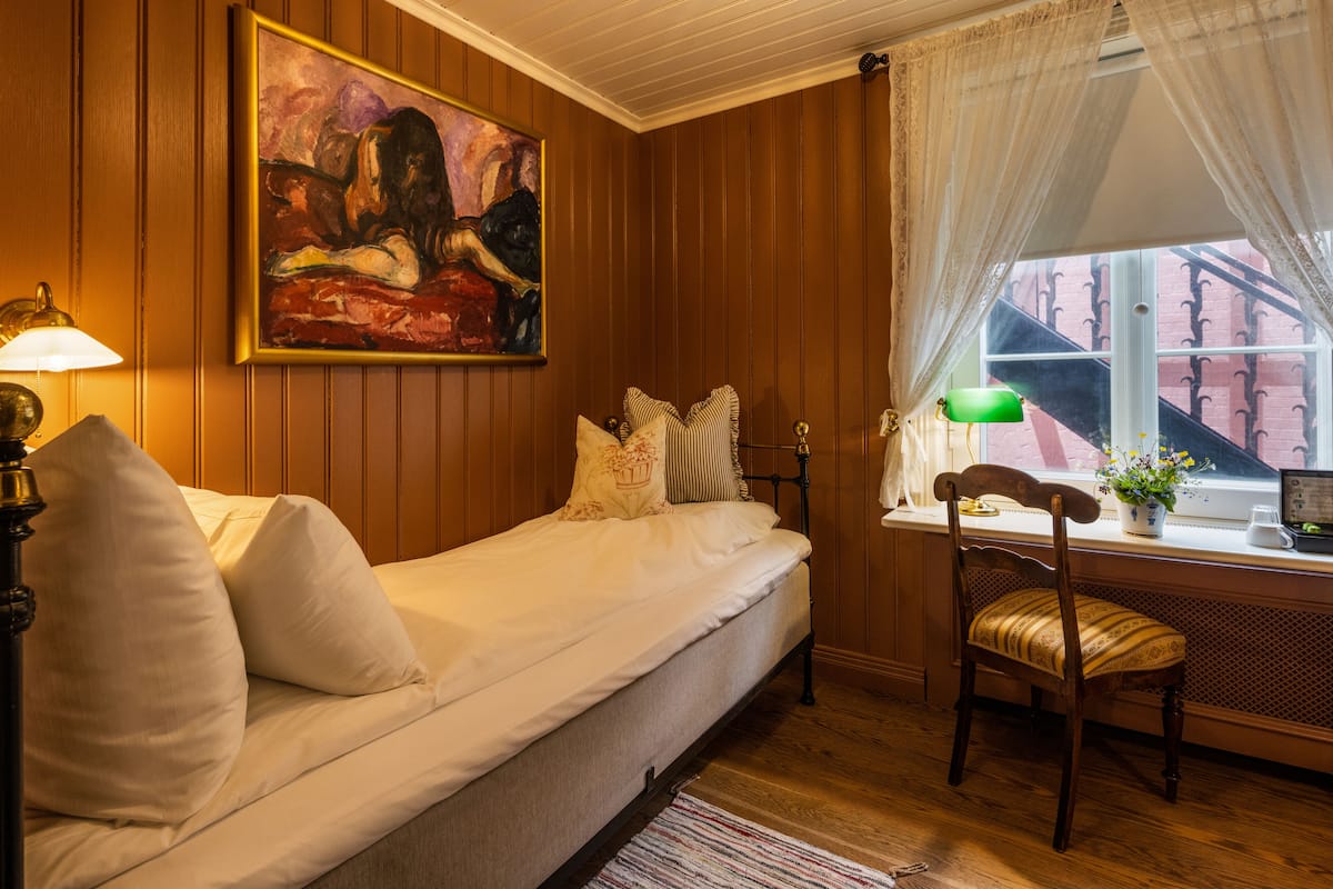 A cozy room with a bed, a chair, a desk with a lamp, a painting on the wall, and a window with curtains.
