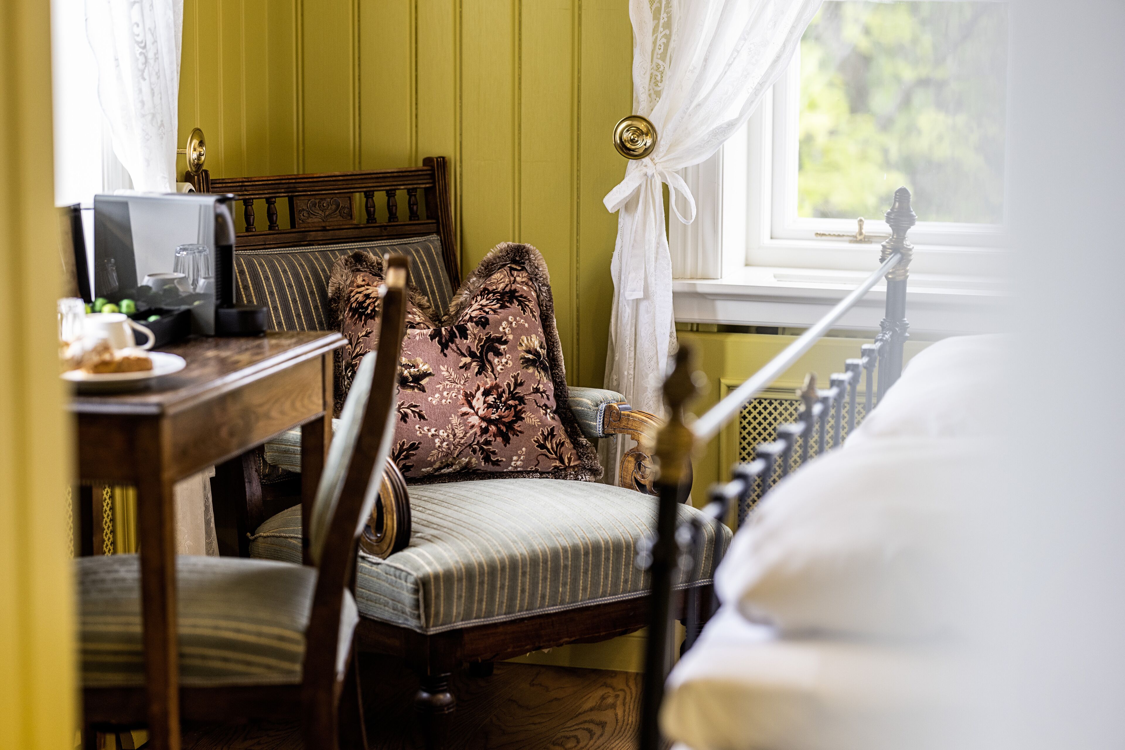 Una habitación con una cama de madera, una silla con un cojín floral, una mesita con una taza y un platillo, y una ventana con cortinas.