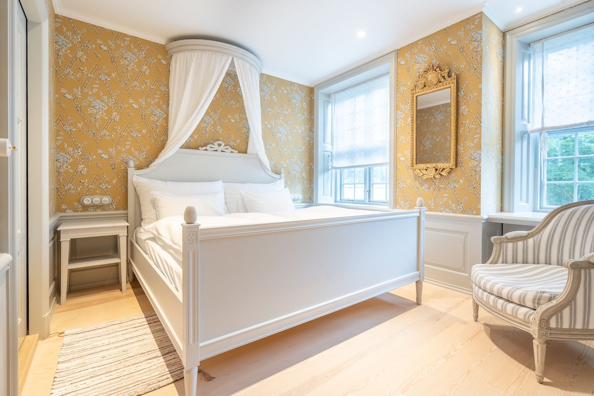 A bedroom with a four-poster bed, a nightstand, a chair, and a window with a view.