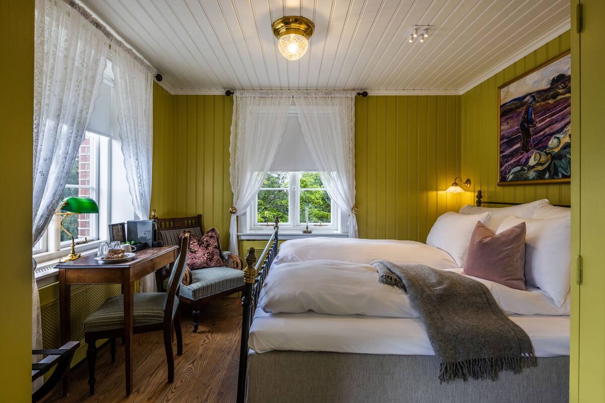 A room with a bed, a desk with a lamp, a chair, and a window with curtains.