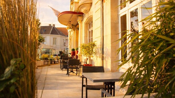 An outdoor cafe with tables and chairs, potted plants, and a building with a balcony. - Logis Hotel Le Majestic (Chatelaillon-Plage)