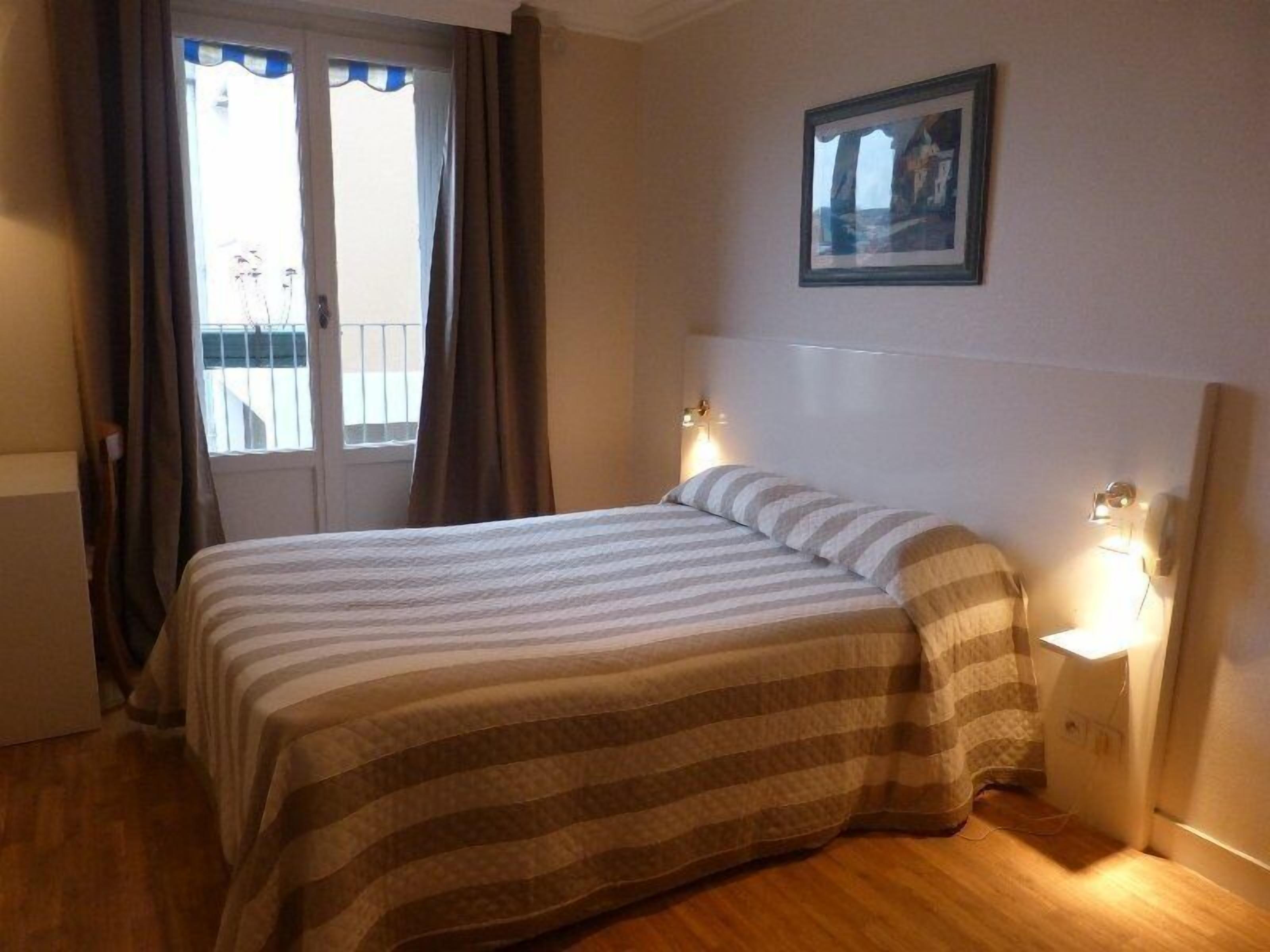 A hotel room with a bed, a window with curtains, a painting on the wall, and bedside lamps.