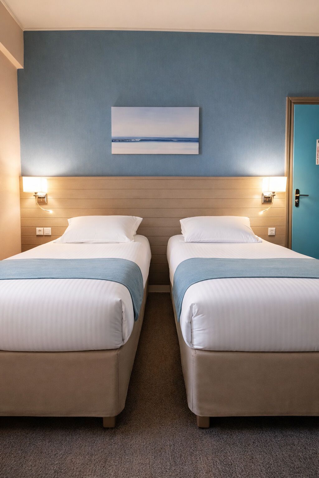 Habitación de hotel con dos camas, una pared azul, un cuadro y una puerta.