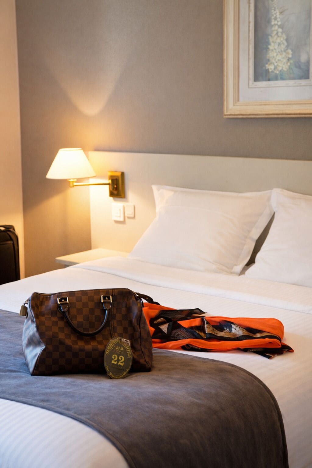 A hotel room with a bed, a lamp, a framed picture on the wall, and a handbag on the bed.