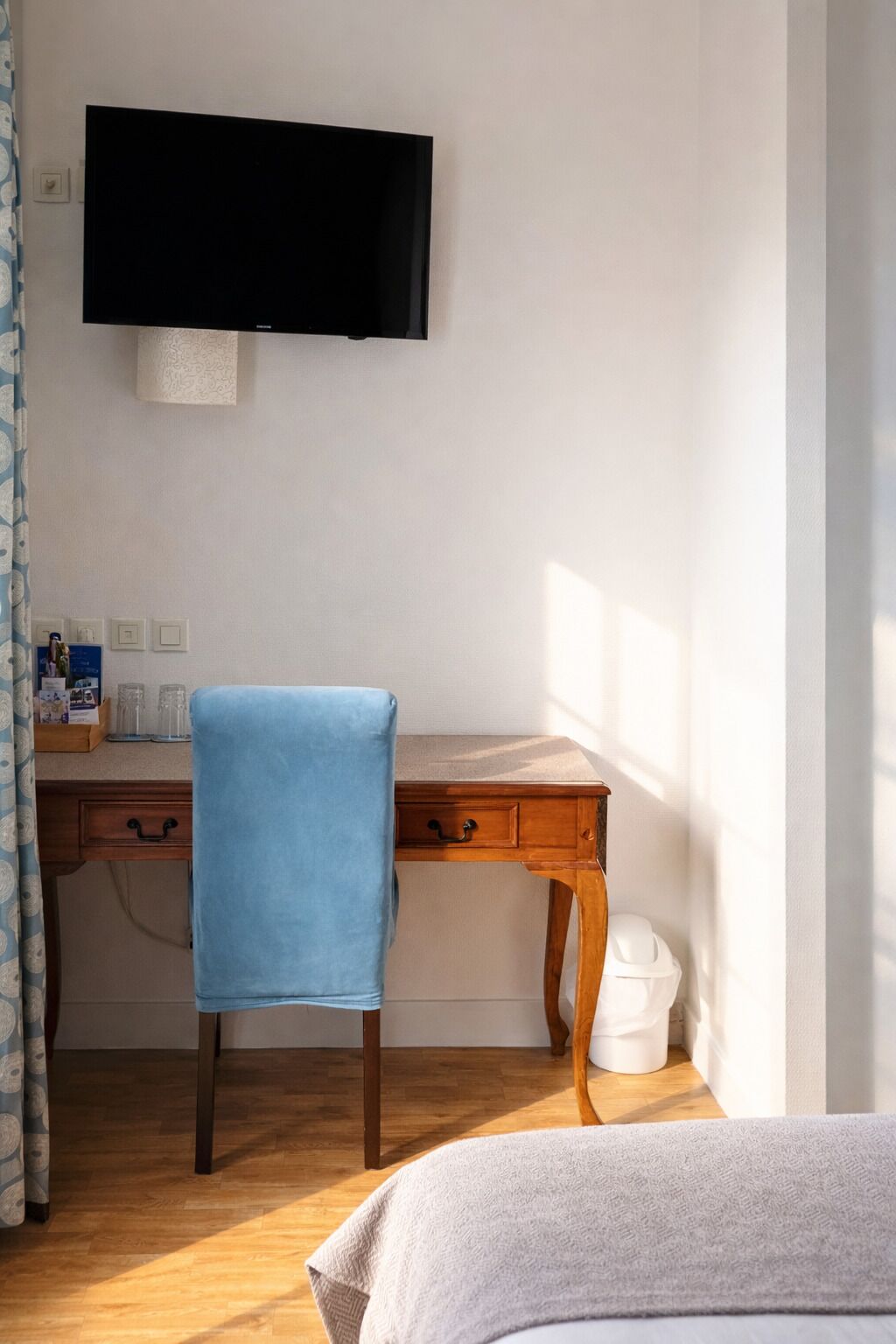 A room with a wooden desk, a blue chair, a television mounted on the wall, and a bed with a grey blanket.