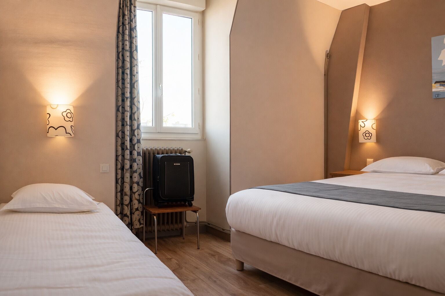 A hotel room with two beds, a radiator, a television on a small table, and wall-mounted lights.