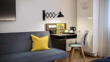 A modern hotel room with a grey sofa, a yellow pillow, a desk with a lamp, a chair, and a wall-mounted phone.