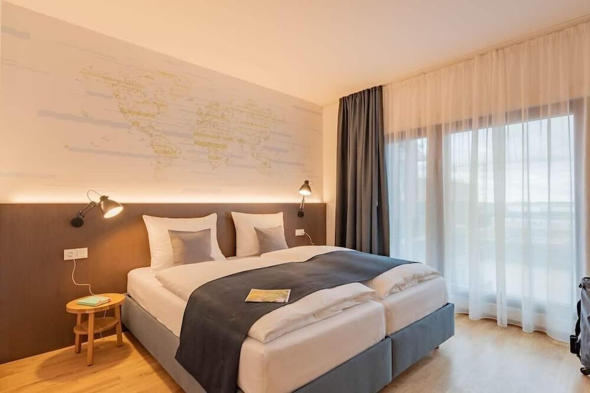Una camera d'albergo con un letto grande, una mappa appesa al muro, un tavolino con una lampada e vista all'esterno attraverso tende leggere.