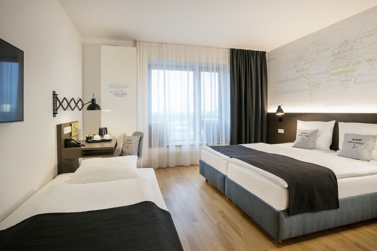 Une chambre d’hôtel avec deux lits, un bureau avec une lampe, une carte accrochée au mur et une vue sur la ville grâce à de grandes fenêtres.