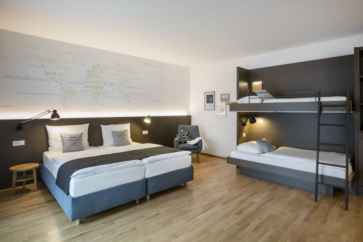 Une chambre d’hôtel moderne avec un grand lit, un lit plus petit, un lit superposé, un bureau et une chaise. Il y a une carte accrochée au mur et une petite table ronde.