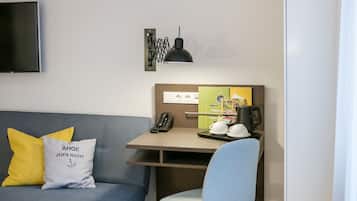 Una habitación de hotel moderna con sofá, escritorio con teléfono y hervidor, una silla y un televisor de pantalla plana montado en la pared.