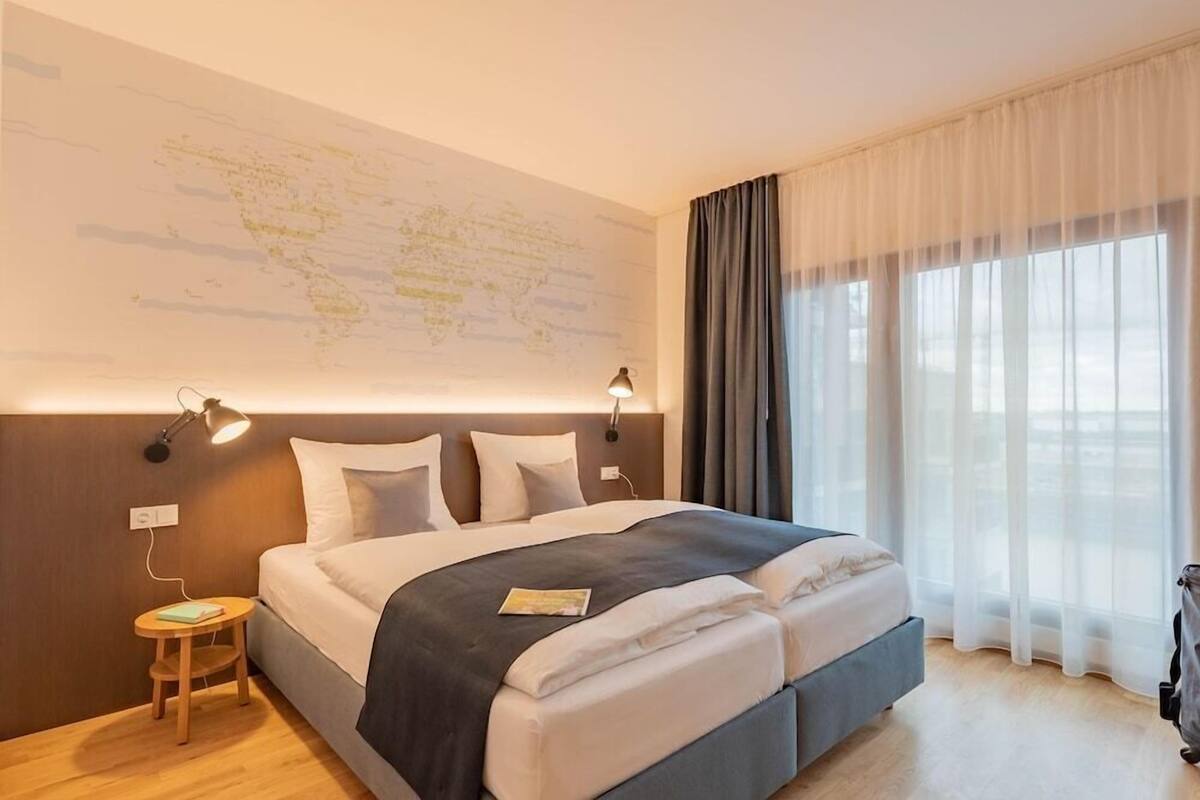 Une chambre d’hôtel avec un grand lit, une carte accrochée au mur, une petite table avec une lampe, et une vue sur l’extérieur à travers de fines rideaux.