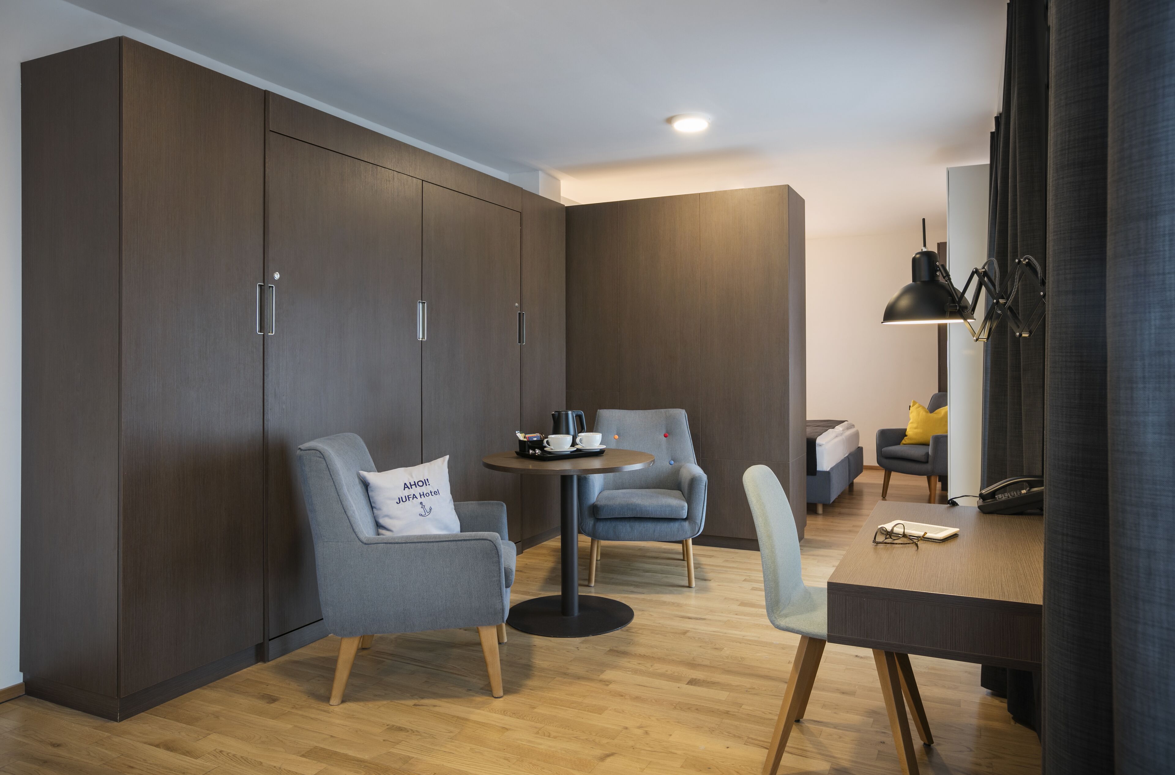 Ein modernes Hotelzimmer mit großem Kleiderschrank, zwei Sesseln, einem kleinen runden Tisch mit Kaffeemaschine und einem Schreibtisch mit Lampe.