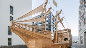 A wooden ship-shaped playground structure with climbing nets and a slide, located in an urban area with modern buildings in the background.
