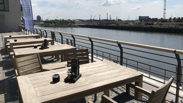 Outdoor wooden tables and chairs by a riverside, with a railing and a view of a cityscape in the background.