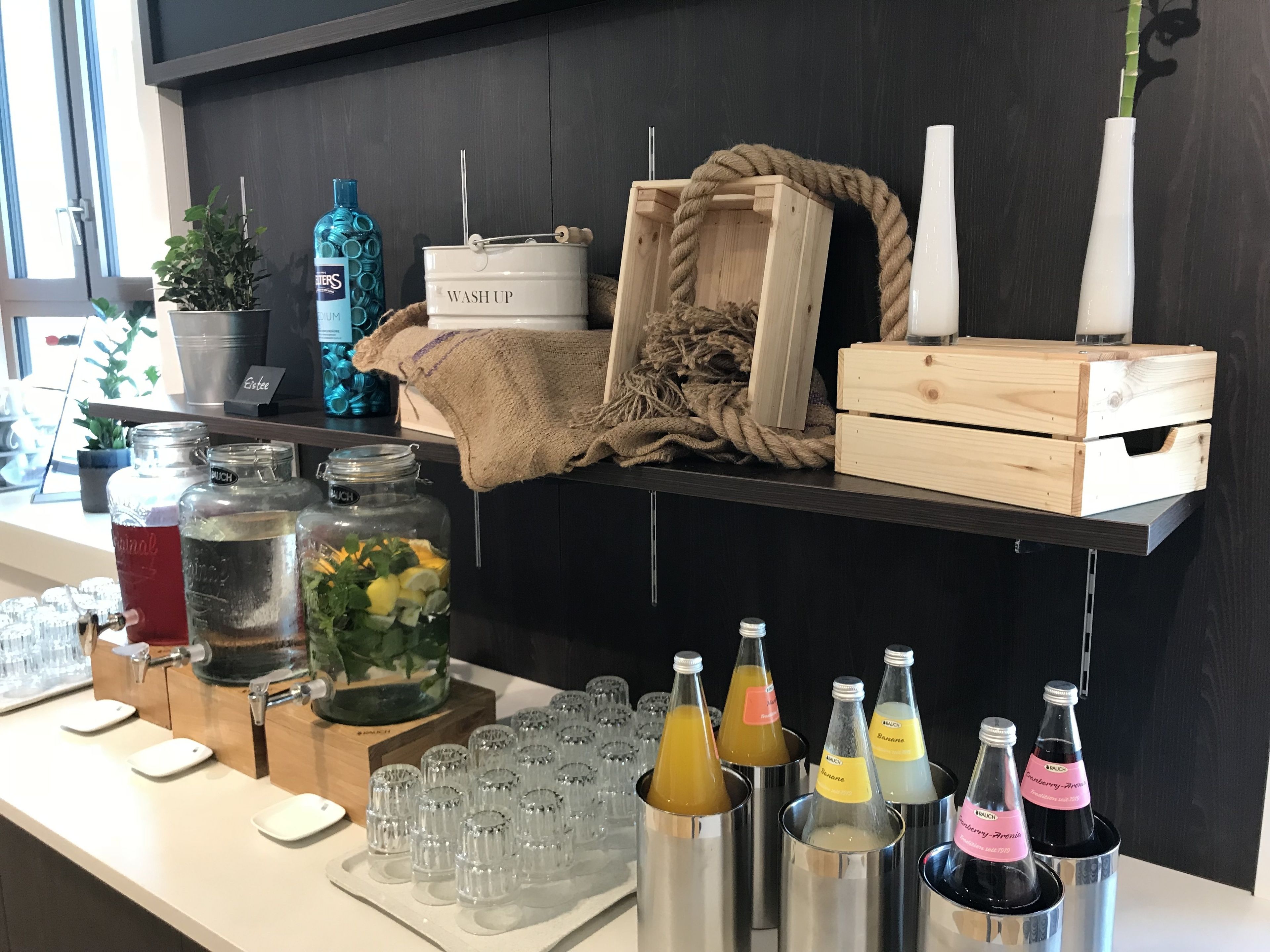 Eine Barbereich mit verschiedenen Getränkeautomaten, einem großen Krug mit Mixgetränken und einem rustikalen Regal mit dem Schild 'WASH UP'.