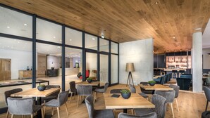 A modern dining area with wooden tables and grey upholstered chairs, large windows, and a visible bar area in the background.