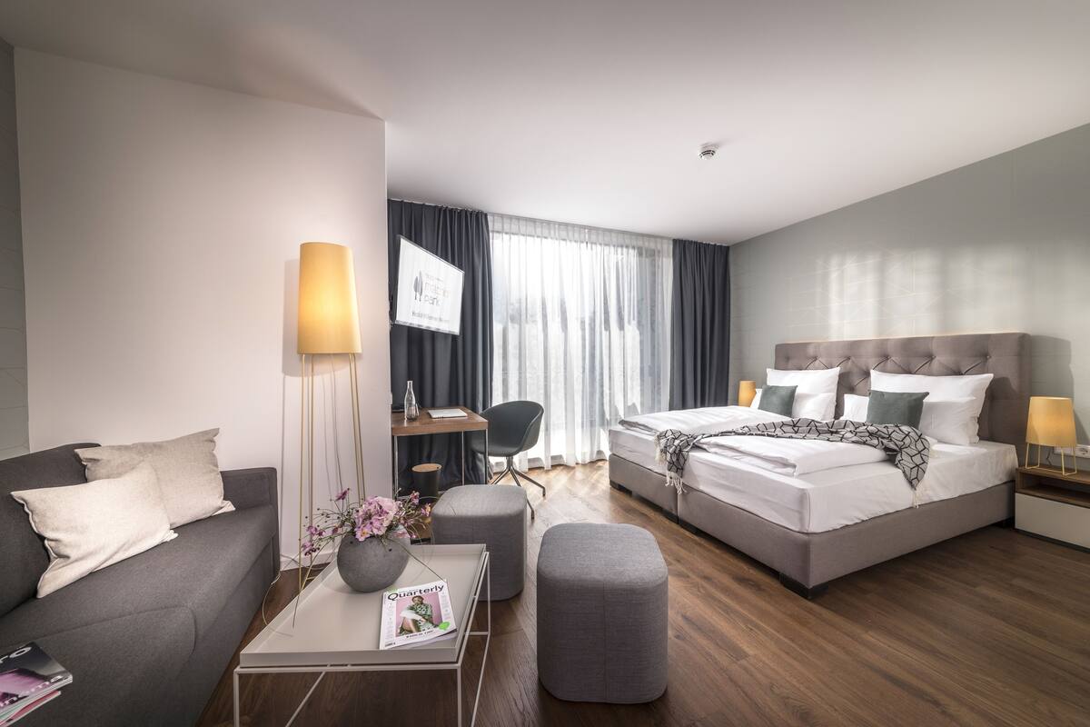 A modern hotel room with a grey sofa, a bed with a grey headboard, a desk with a computer, a chair, and a coffee table with a magazine.